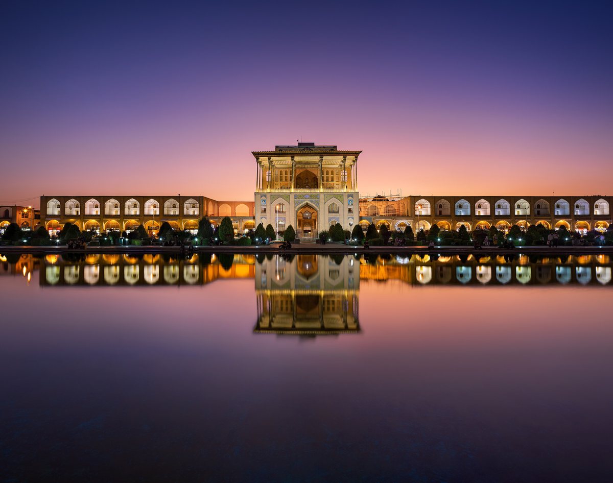 Ali Qapu Palace and the arcades of Naqsh-e Jahan Square lit at dusk, reflected in the square's long pool.
