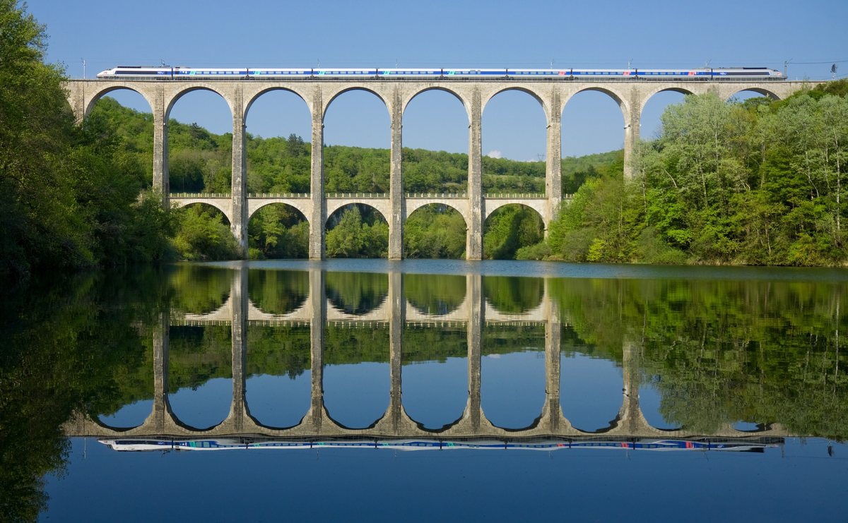A high-speed train crossing the two tiers of stone arches of the Cize–Bolozon viaduct, mirrored in the still river below.