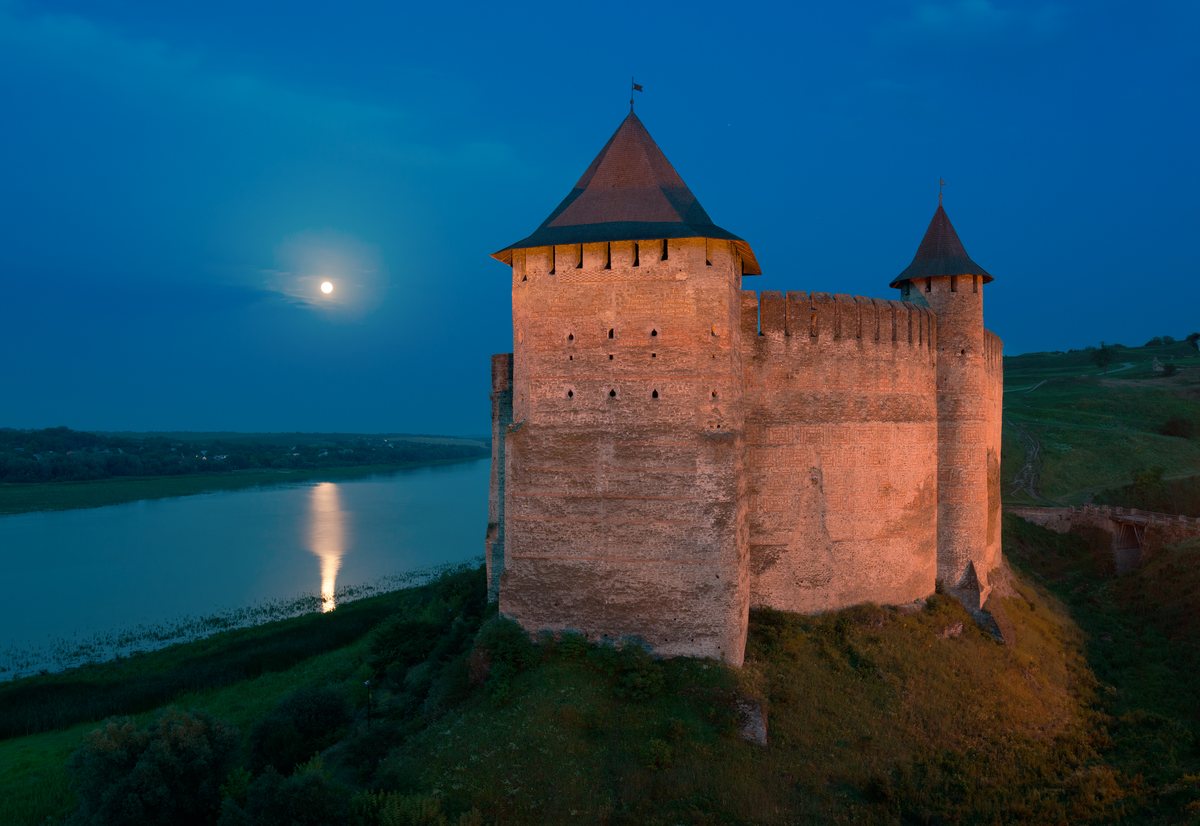 The towers of Khotyn Fortress lit warm at dusk above the Dniester, with the full moon reflected in the river.