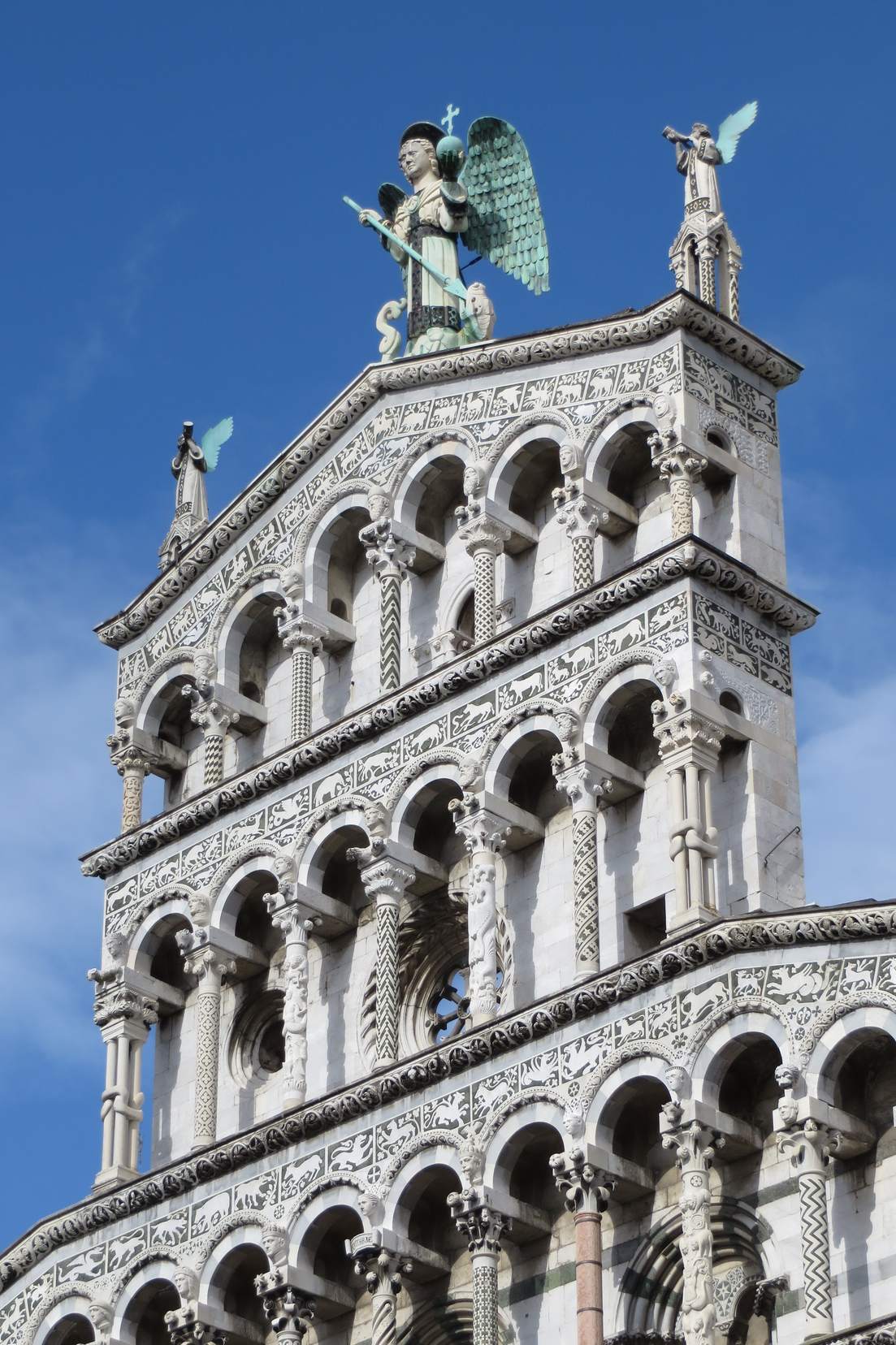 The arcaded marble facade of San Michele in Foro in Lucca, topped by a statue of the Archangel Michael, against a blue sky.