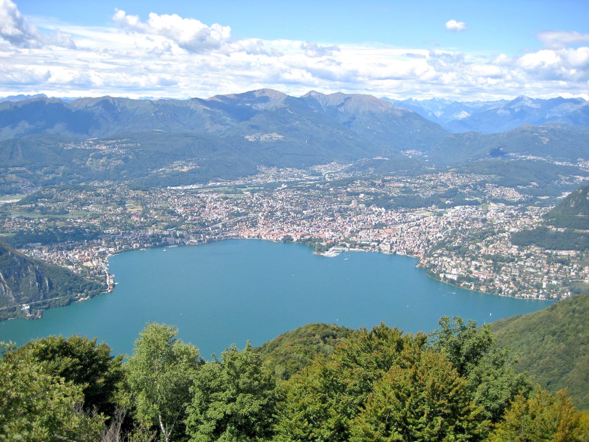 Lake Lugano and the city of Lugano spread below mountains, seen from the summit of the Sighignola.